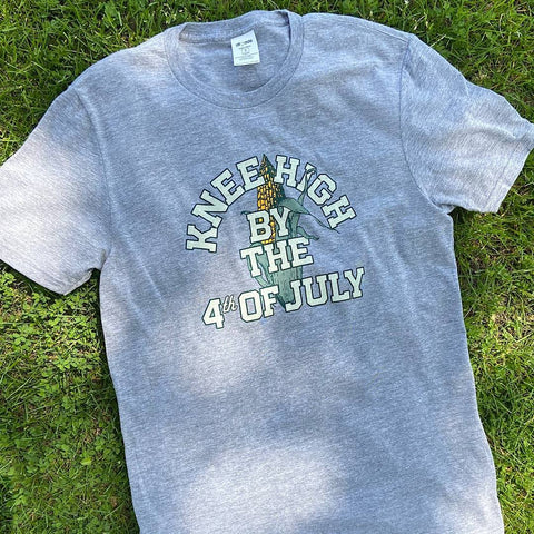 gray T-shirt on grass with words “knee high by the 4th of July” on top of single cob of corn